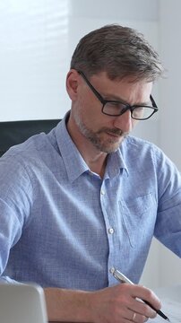 Businessman wearing blue sleek shirt, glasses, is thinking and concentrating while working at the desk in a bright, modern office, demonstrating professionalism and efficiency. Business people concept