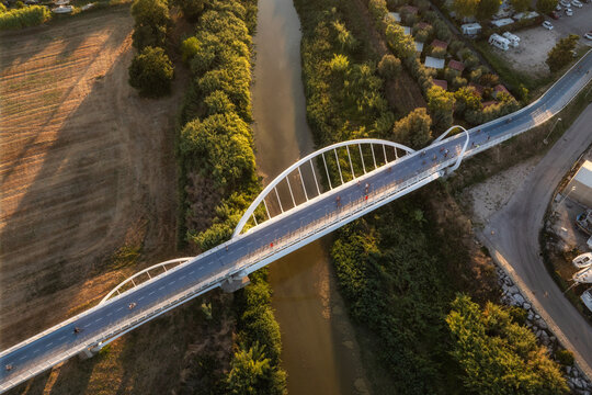 Aerial view of a modern white arch bridge crossing a river with cyclists riding across, surrounded by lush greenery and fields in Porto San Giorgio, Marche, Italy.