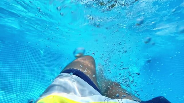 Point of view underwater shot of a man in swim trunks kicking and floating in a blue swimming pool with rising air bubbles. Summer vacation video.
