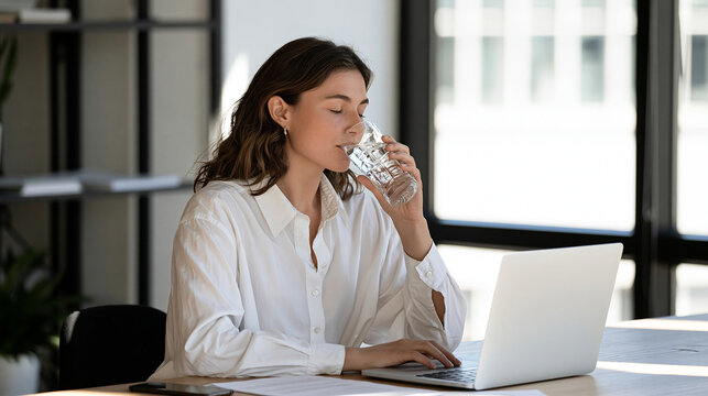 Woman drinking water while working at a computer desk, healthy work routine, workplace hydration, employee wellness, productivity, healthy office lifestyle, remote work, natural