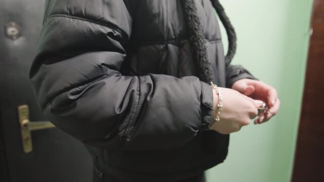Closeup white woman hands holding keys against black puffer jacket, slow fumbling motion to unlock metal door, visible bracelet and keyring, tense body language in narrow corridor, realistic urban