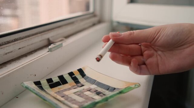 Hand holding cigarette on windowsill, freelance writer taking quiet pause, glass ashtray on sill, soft morning light, blurred city skyline, ember glowing, intimate domestic mood and reflective