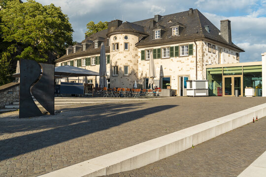 View of the historic building at Drachenfels plateau with outdoor seating, modern glass extension, and abstract metal sculpture on a paved square in Konigswinter, North Rhine-Westphalia, Germany.