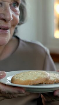 Vertical video: Bringing white plate, senior woman offering two cookies at home to off-camera guest