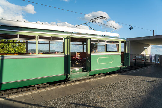 View of the Drachenfelsbahn rack railway train at the station, with green carriage, open doors, and cobblestone platform under a blue sky in Konigswinter, North Rhine-Westphalia, Germany.