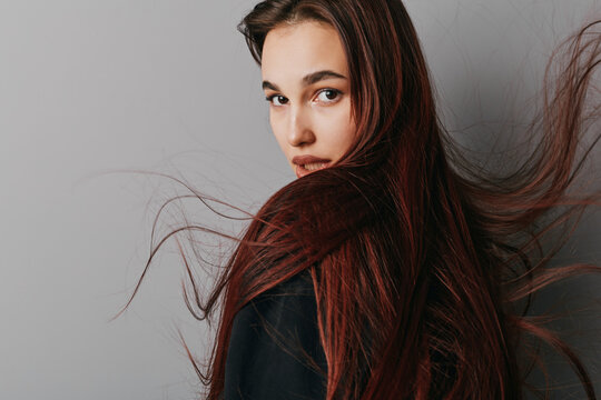 Young woman with long brown hair and serious expression looking over shoulder against plain background. Closeup portrait with natural makeup and casual black outfit.