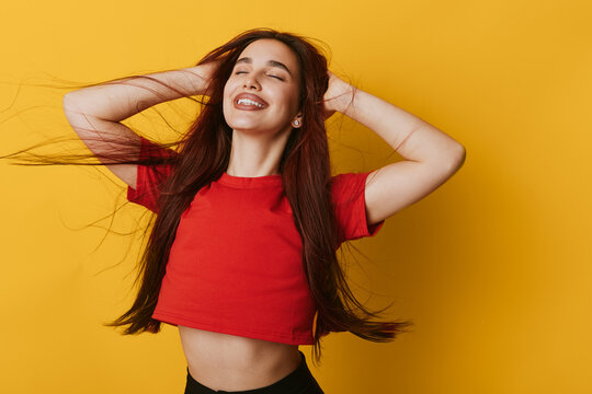 Young woman with long hair smiling and posing with hands in hair wearing red crop top against yellow background. Happy female model expressing joy and confidence in studio.