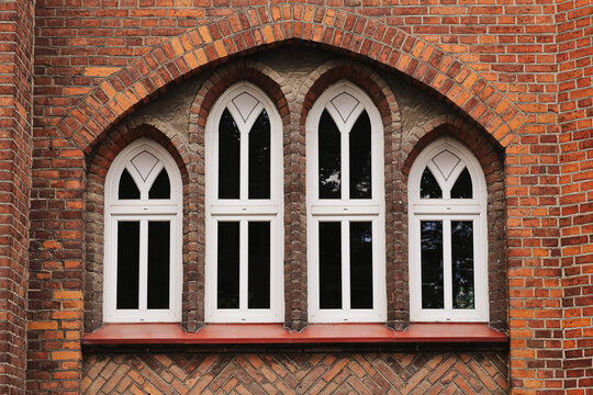 Gothic windows decorate brick facade. White frames contrast red masonry. Historic arches have pointed shapes. Brickwork surrounds symmetrical openings. Old building forms architectural background.