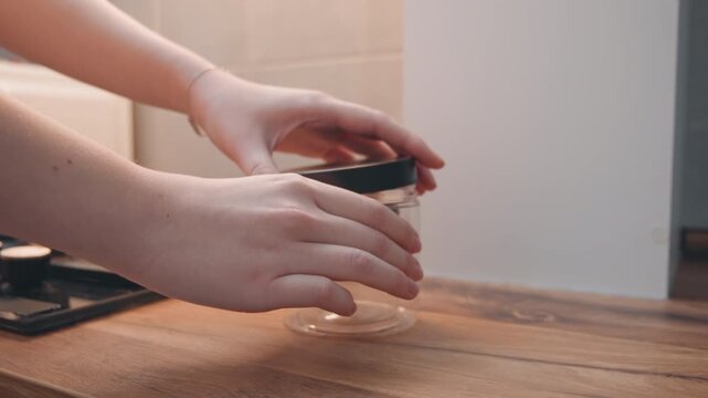 Wood counter with jar and hands preparing. Calm home environment with focused food preparation details. Closeup of hands gently handling jar in peaceful kitchen setting with natural light