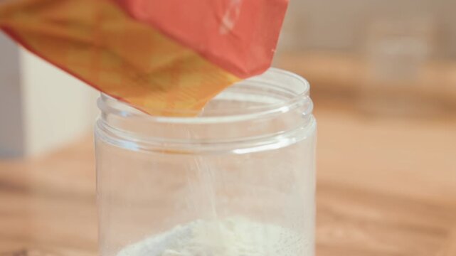 Warm natural light highlights pouring flour. Home baker sifting flour into jar in cozy pantry setting. Closeup of flour gently pouring into jar amid warm natural lighting and rustic ambiance