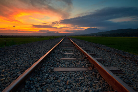 View of railway tracks stretching through green fields towards mountains under a vibrant sunset sky in Asinan, Bawen, Central Java, Indonesia.