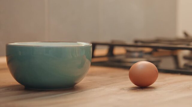 Empty bowl waits beside brown egg, ceramic bowl on wooden counter, hand reaches into frame, gas stove blurred in background, warm natural light, calm countertop staging for recipe