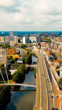 Vertical video. Ghent, Belgium. Esco (Scheldt) river embankment, sweeping aerial panorama over historic rooftops, modern avenues and tree-lined waterways under a cloudy summer sky. Drone footage. Ric