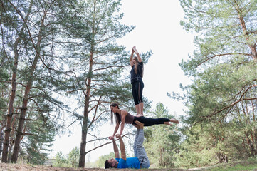 Fototapeta premium Man supports two women performing a challenging three person acro yoga tower with plank and standing balance outdoors in a sunny pine forest.