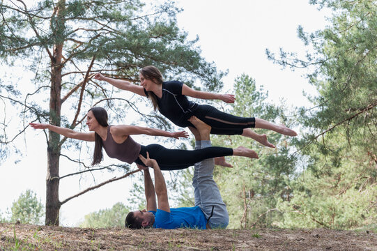 Man supports two women in horizontal flying acro yoga poses, demonstrating strength, trust and coordinated balance outdoors in a pine forest.