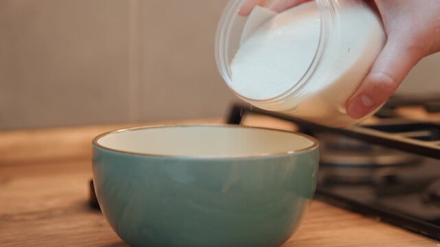Pouring sugar into teal mixing bowl, steady hand measuring granulated sugar from jar, grainy cascade of crystals, ceramic bowl on wooden counter, baker preparing batter for dessert, warm studio light
