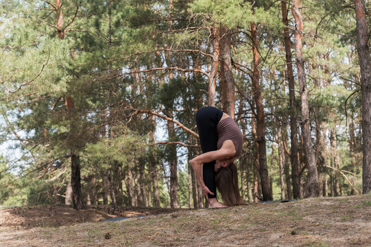 Flexible athletic woman performs a deep standing forward bend, stretching her legs and back on a yoga mat outdoors among tall pine trees in a forest.