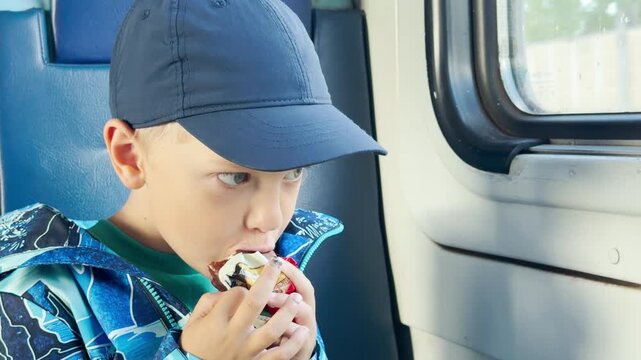 A happy young boy happily eats a sweet snack while sitting on a blue train seat. He looks forward with a joyful and curious expression during his cozy journey.