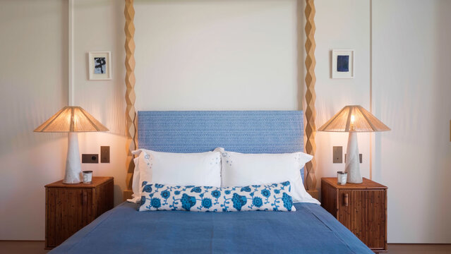 A hotel bedroom featuring a blue upholstered bed with a patterned bolster pillow, wooden bedside tables, and rattan-shaded lamps