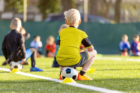 Young Soccer Player Sitting on Football During Practice Break - Kids Training Session on Green Grass Pitch with Yellow Cones