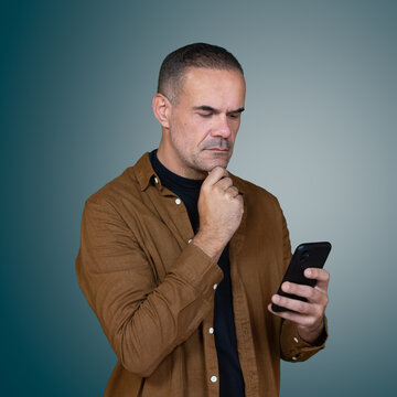 Thoughtful middle aged man looking at smartphone with hand on chin, studio portrait representing decision making, online research and mobile technology.