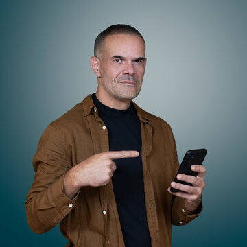 Confident middle aged man pointing at smartphone while looking at camera, studio portrait with copy space for mobile technology and digital marketing.