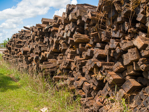Massive stack of heavy old wooden railroad ties featuring rusty metal end plates lying on the green grass next to the railway tracks.