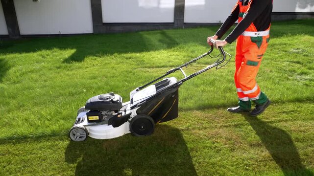 Worker mows vibrant green lawn with gas-powered mower. Bright orange uniform stands out against fresh grass. Focused effort shows in steady, deliberate pushing motion