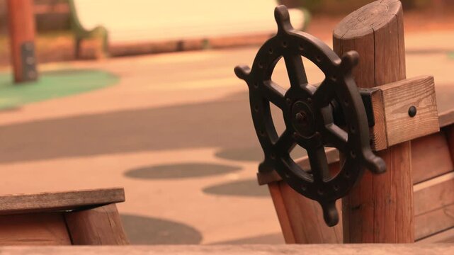 Black metal pirate ship steering wheel mounted on wooden post, playful nautical helm on childrens playground boat structure, imaginative adventure play area outdoors, empty scene, no children present