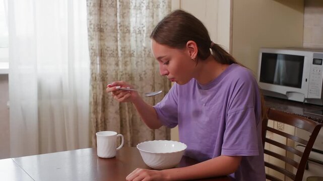 Young woman in purple shirt eating cereal from white bowl at dining table during morning routine in home kitchen with microwave visible.