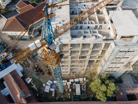 Aerial view of a construction site featuring a large tower crane, unfinished concrete building structure with scaffolding, and surrounding buildings in Banyumas Regency, Central Java, Indonesia.