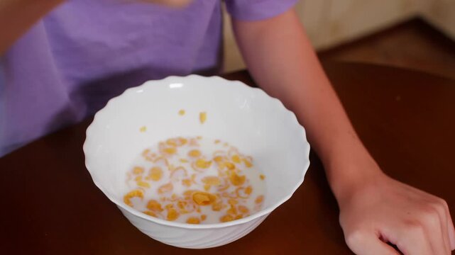 Person in purple shirt holds white bowl of golden cereal with milk while eating breakfast using spoon at table.