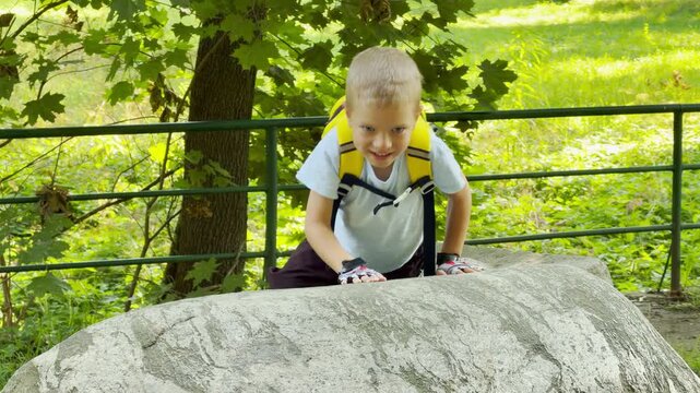 A happy young boy wearing a yellow backpack climbs a large rock. He feels excited and playful during his joyful outdoor adventure in a sunny green park.