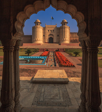 View of Lahore Fort framed by an ornate archway, featuring manicured gardens with vibrant flowers, historic stone walls, and a clear blue sky in Lahore, Punjab, Pakistan.