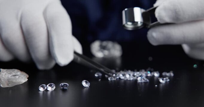 Slow motion close up jeweler sorting loose diamonds on black tabletop with loupe and tweezers.