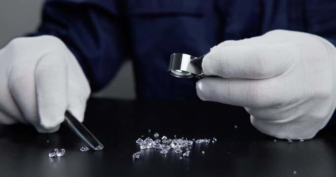 Tracking shot of jeweler picking and inspecting loose diamonds on black table with loupe and white gloves.