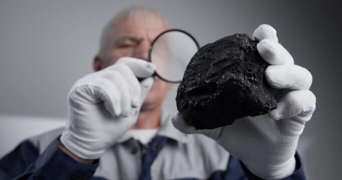 Handheld close up of man examining coal specimen in laboratory with magnifying glass and white gloves.