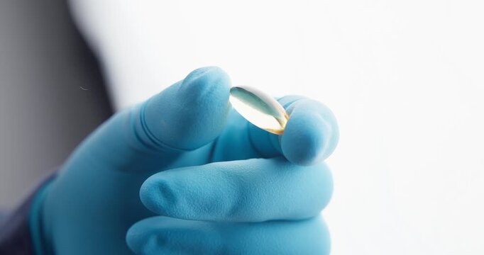 Gloved hand holding softgel capsule with blurred person background.
