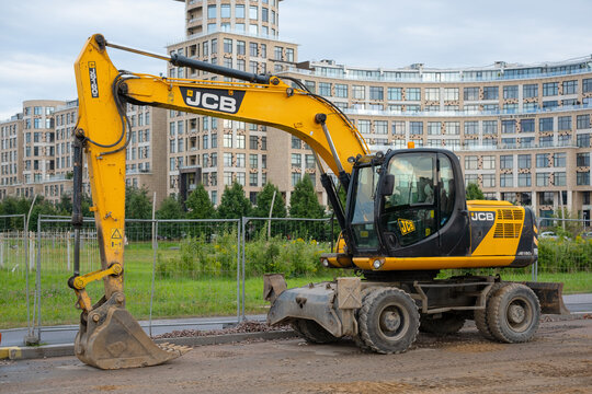 SAINT PETERSBURG, RUSSIA - AUGUST 16, 2026: A yellow JCB JS160W wheeled excavator stands parked on a dirt road near a fenced construction area, with modern residential apartment buildings 