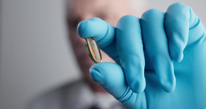Closeup gloved fingers holding softgel capsule white background.