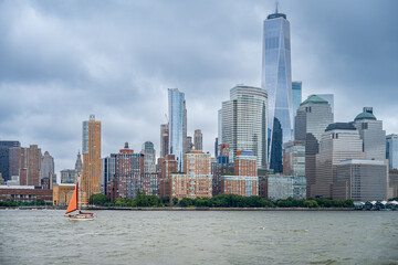 Obraz premium Sailboat passing Lower Manhattan and One World Trade Center. Wide river view shows an orange sail beneath clustered towers and a gray overcast sky.