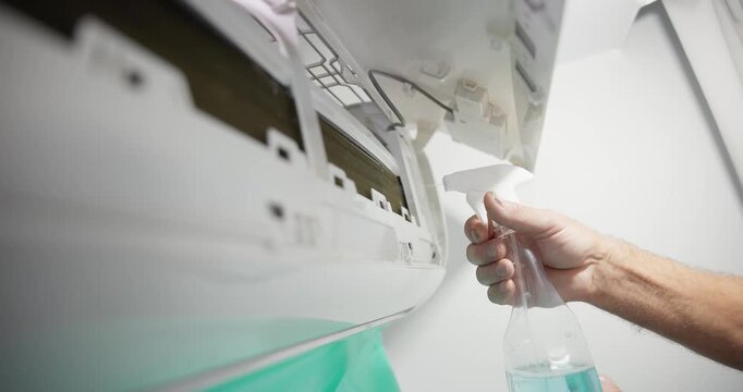 Hand spraying cleaner into indoor air conditioner unit on wall during maintenance.