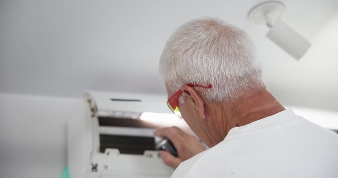 Tracking shot older man inspecting and repairing wall mounted air conditioner inside residential interior during daytime.