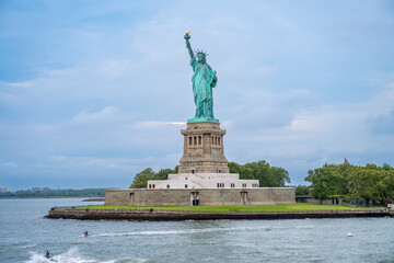 Statue of Liberty standing on pedestal near New York harbor. Green copper landmark on stone base with water splashes and overcast blue sky in background. © Vadim