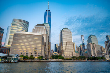 Naklejka premium One World Trade Center above Battery Park City at blue hour. Wide waterfront view shows office towers with lit windows, rippled river water and a blue evening sky.