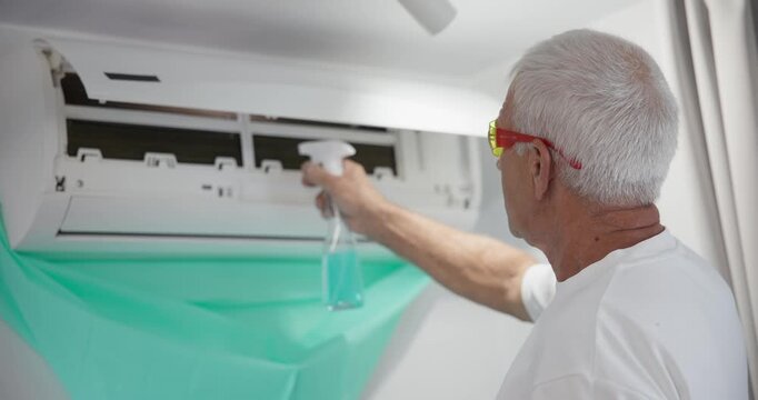 Senior technician cleaning wall mounted air conditioner indoors using spray bottle and protective sheet.