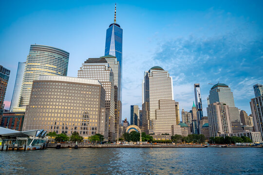 One World Trade Center above Battery Park City at blue hour. Wide waterfront view shows office towers with lit windows, rippled river water and a blue evening sky.
