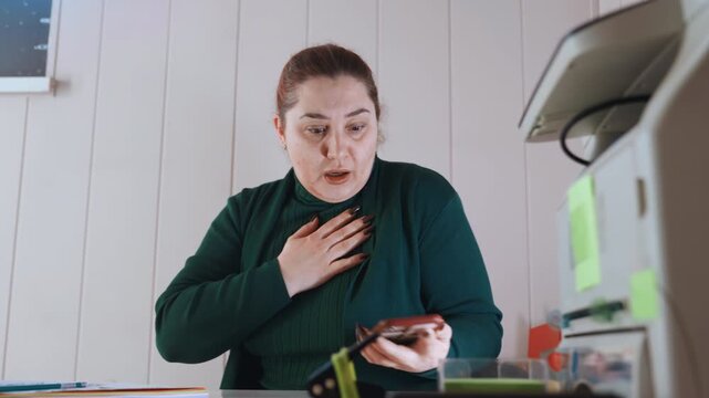 Woman alerting dispatcher about chest pain, hand on sternum, glances toward window and door, tense posture at home desk, alternating between phone and note taking while awaiting paramedic response