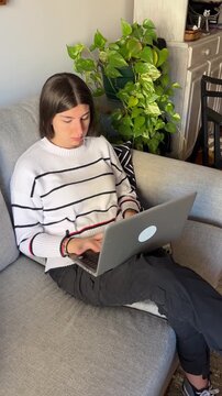 Young woman focused on working or studying on her laptop from home, sitting on a comfortable sofa in a cozy living room.