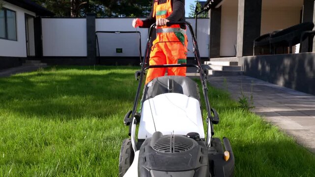 Worker pushes mower across green lawn. Gloved hands grip handle, guiding machine. Bright orange suit stands out against grass. Sunlight casts clear shadows on paved path